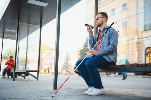 Jeune aveugle assis sur le banc d'un abribus, tenant une canne et étant en appel avec son smartphone